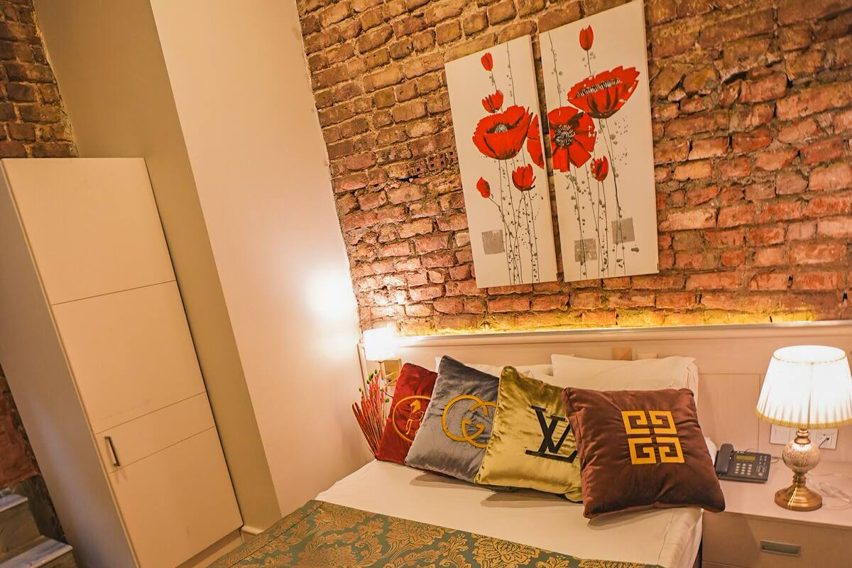 A bedroom with a brick wall, a bed with patterned bedding, a nightstand with a lamp, and a wardrobe.