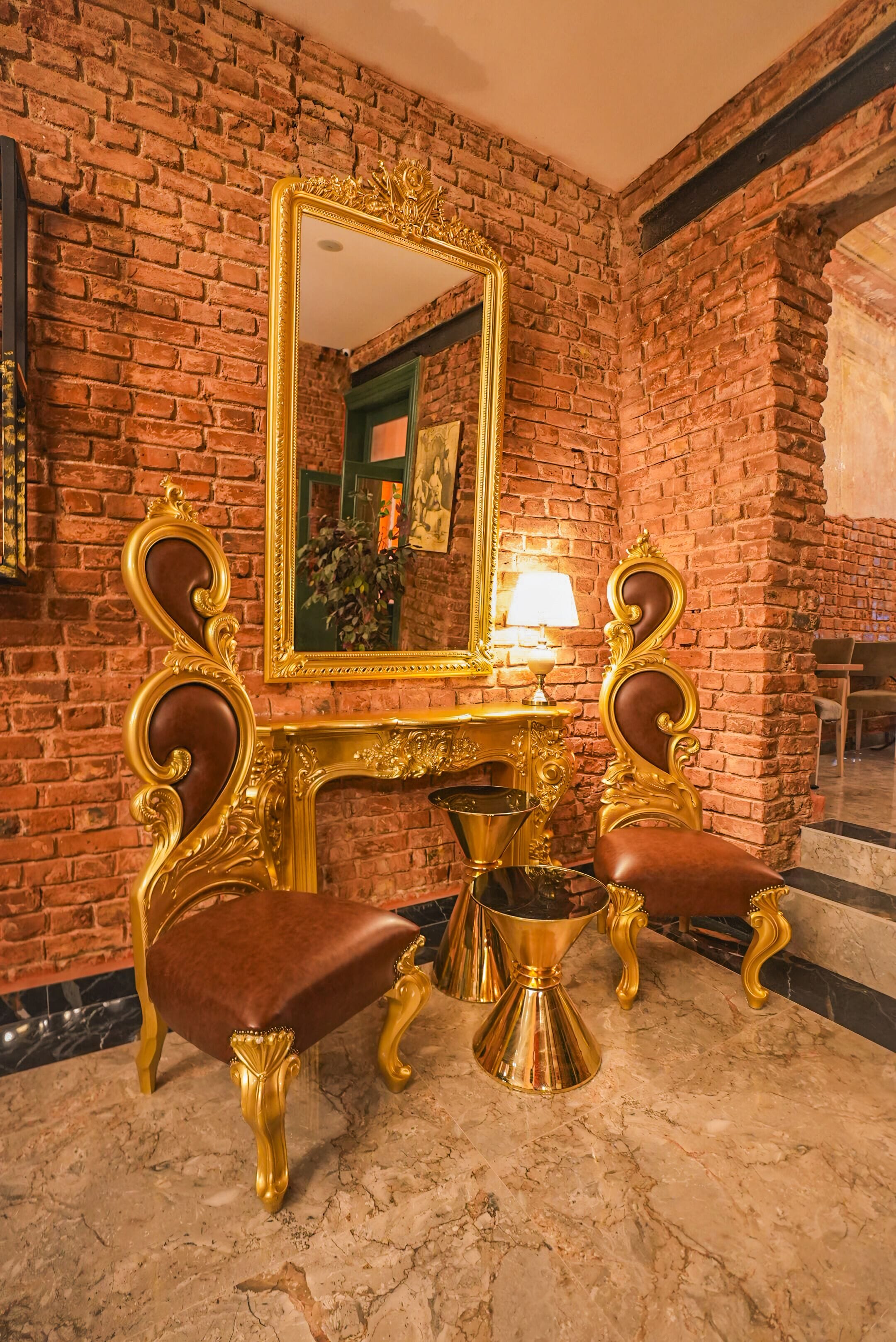 A room with a brick wall, a gold-framed mirror, a marble floor, and two upholstered chairs with gold legs.