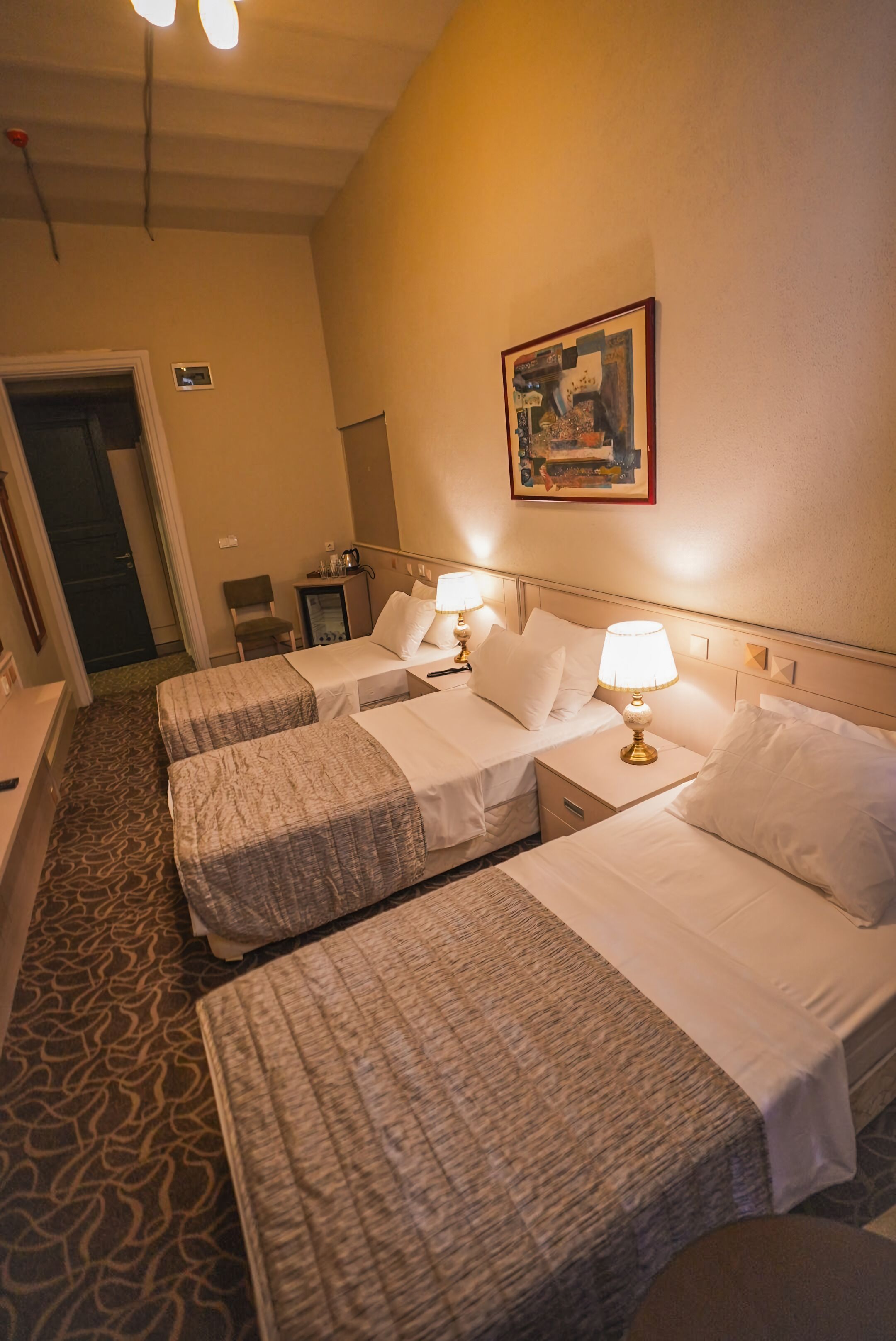 Habitación de hotel con dos camas, cada una con mesita de noche y lámpara, una silla, un escritorio y una puerta.