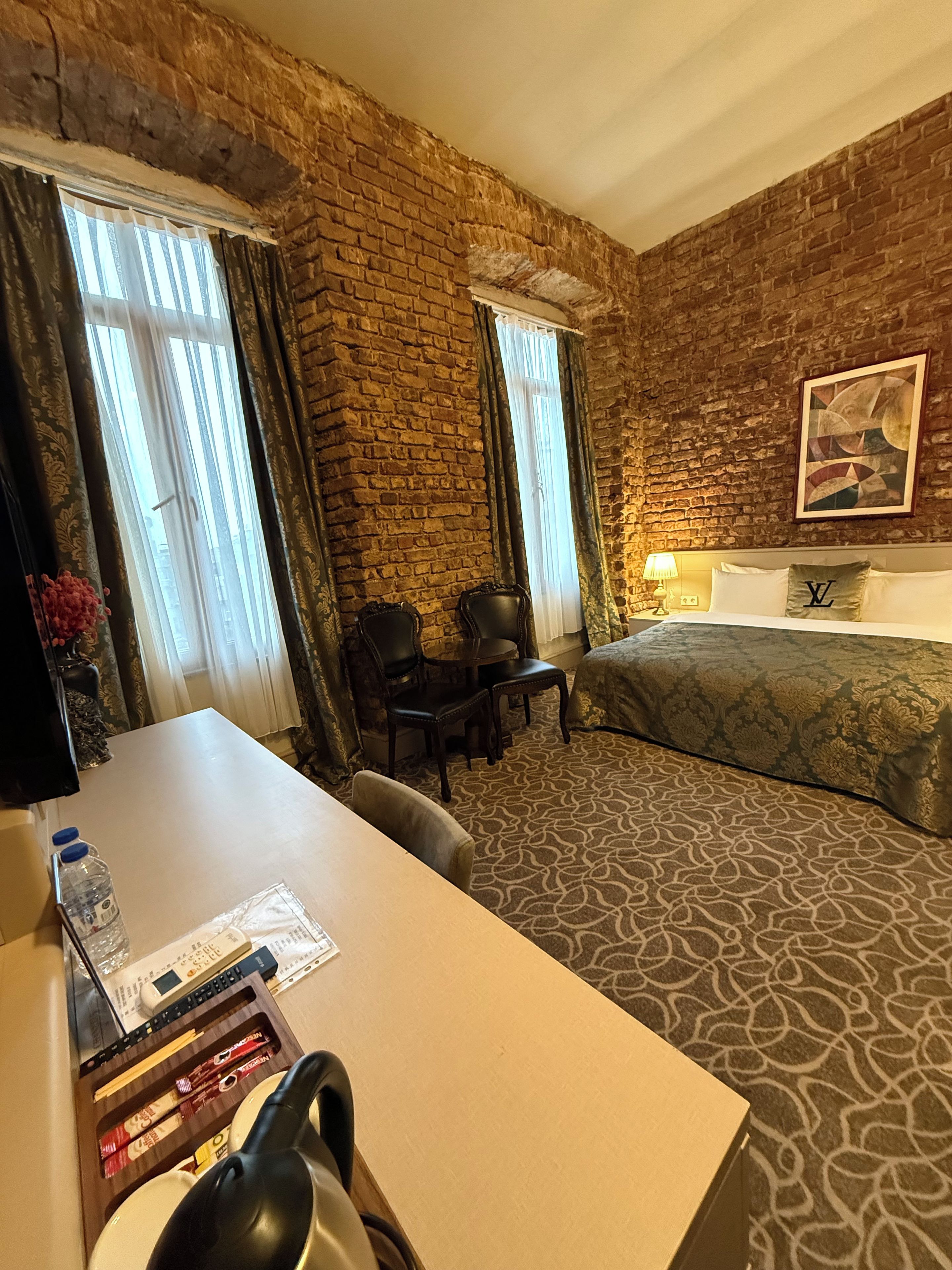 A hotel room with a brick wall, a bed, a desk with a chair, a table with a chair, and a lamp.