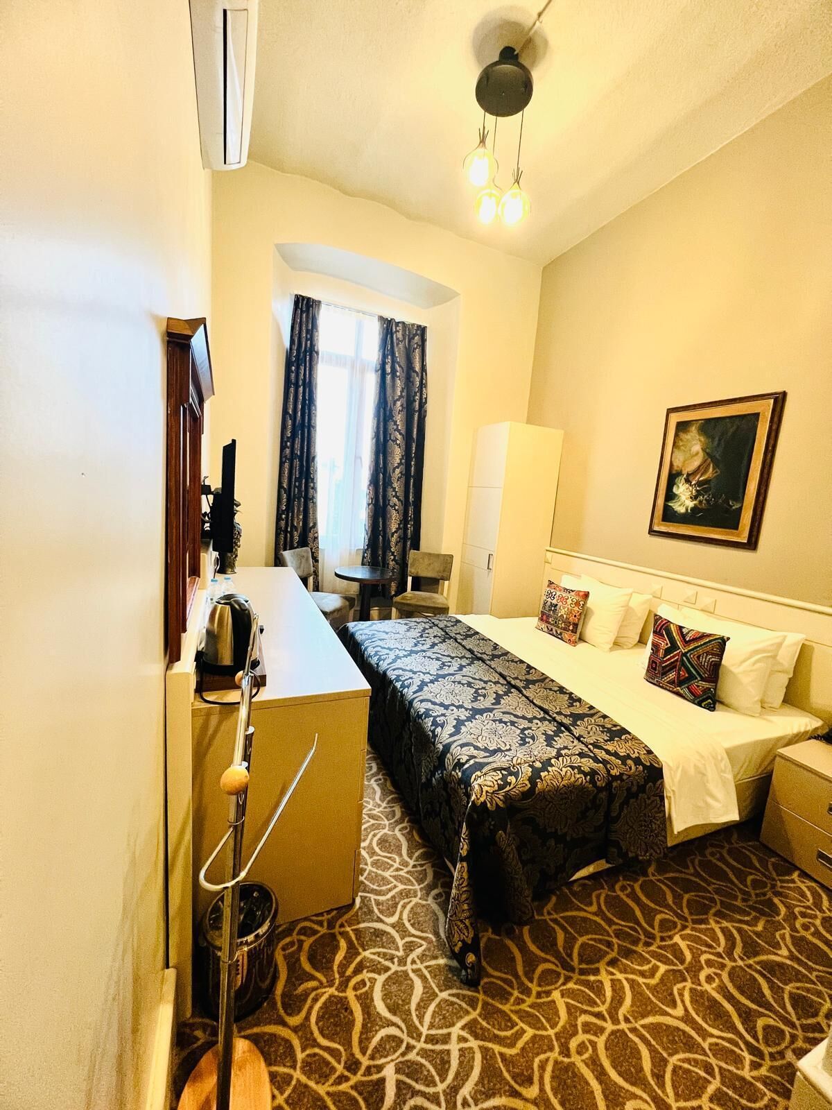 A hotel room with a bed, a desk, a chair, a wardrobe, and a painting on the wall.