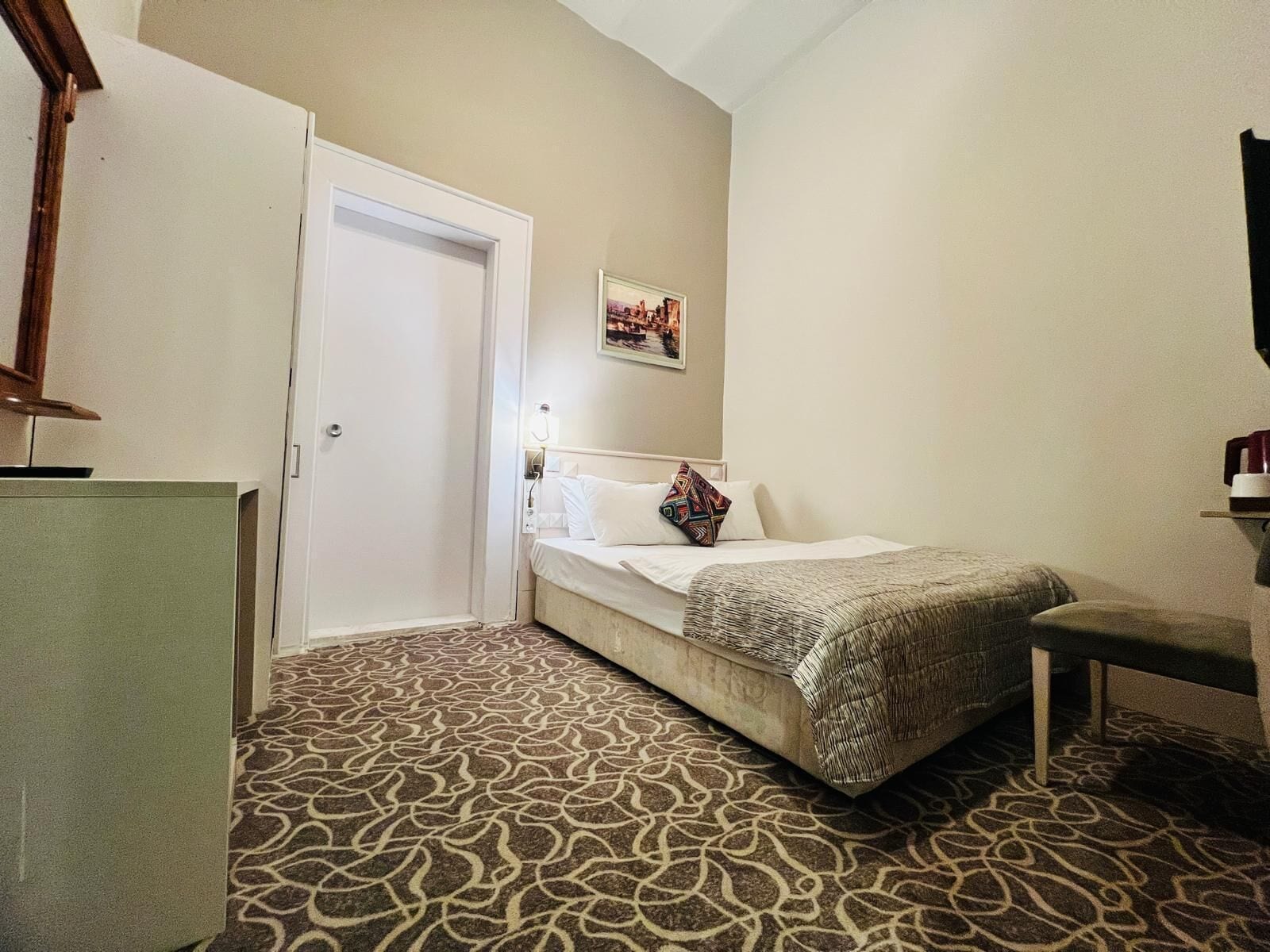 A hotel room with a bed, a desk, a chair, a door, and a patterned carpet.