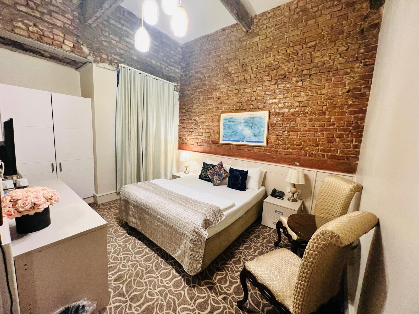 A hotel room with a brick wall, a bed with multiple pillows, a desk with a computer and flowers, and a chair.