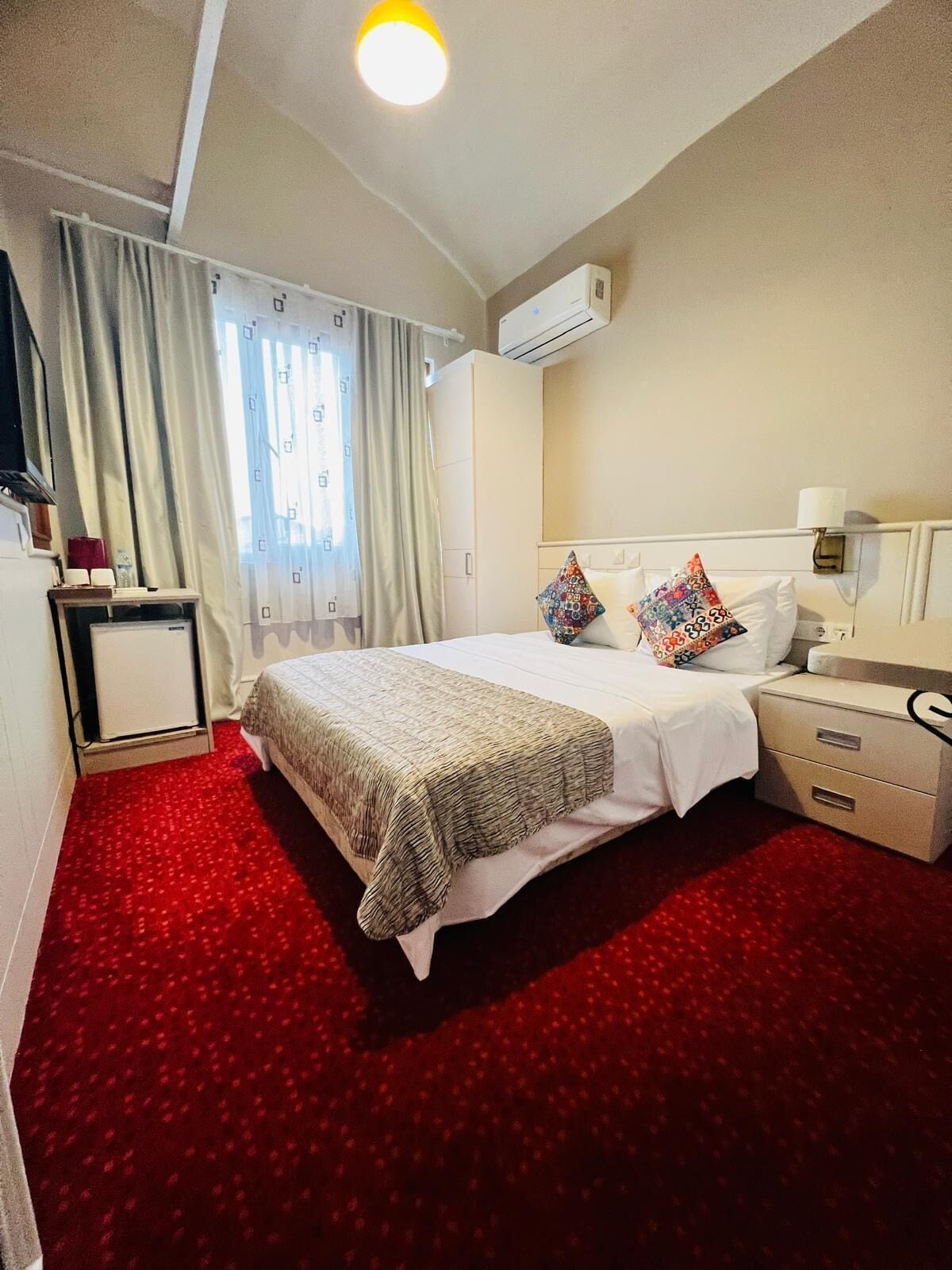 A hotel room with a large bed, a bedside table, an air conditioning unit, and a red carpet.