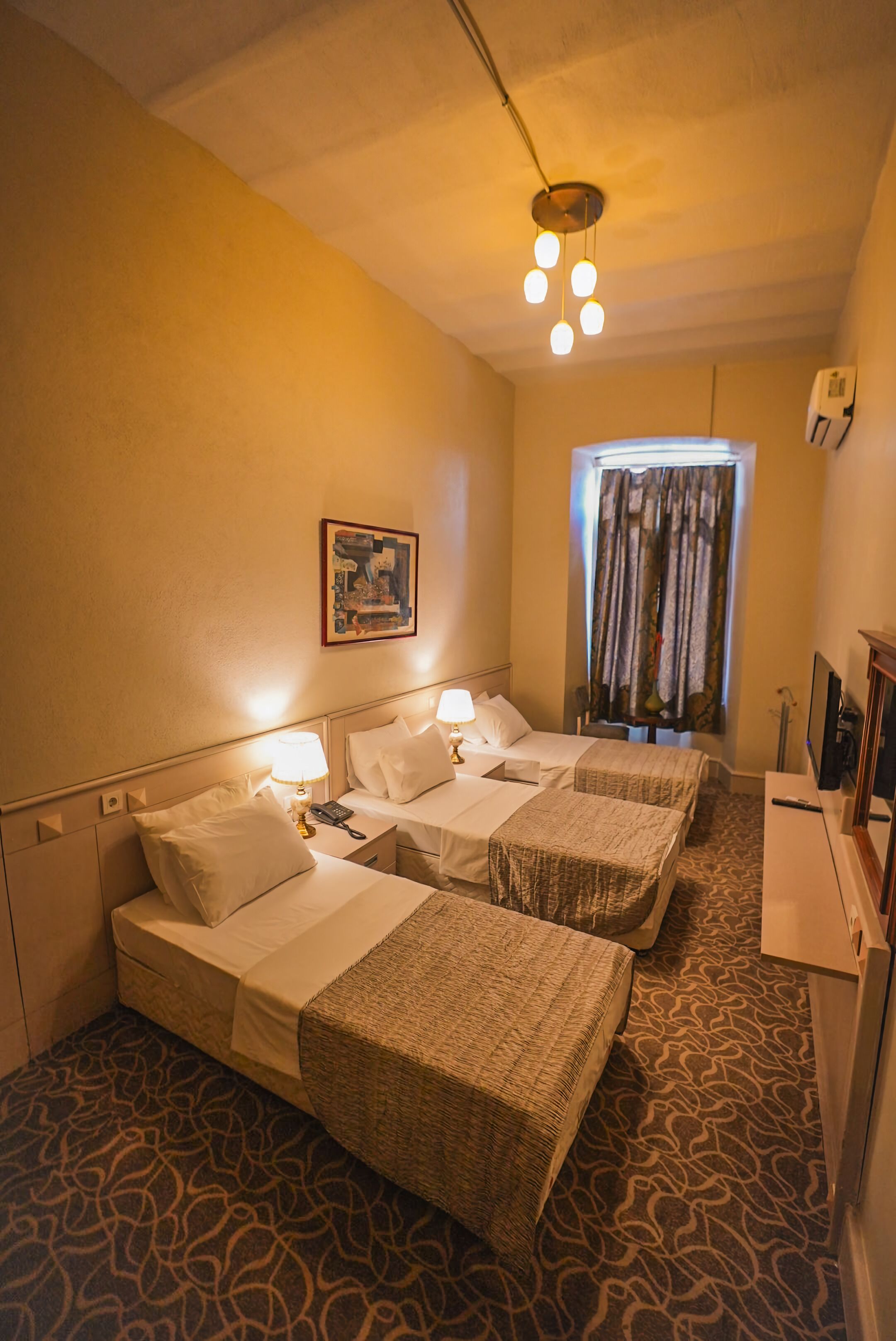 A hotel room with two beds, a desk, a lamp, a painting on the wall, and a window with curtains.