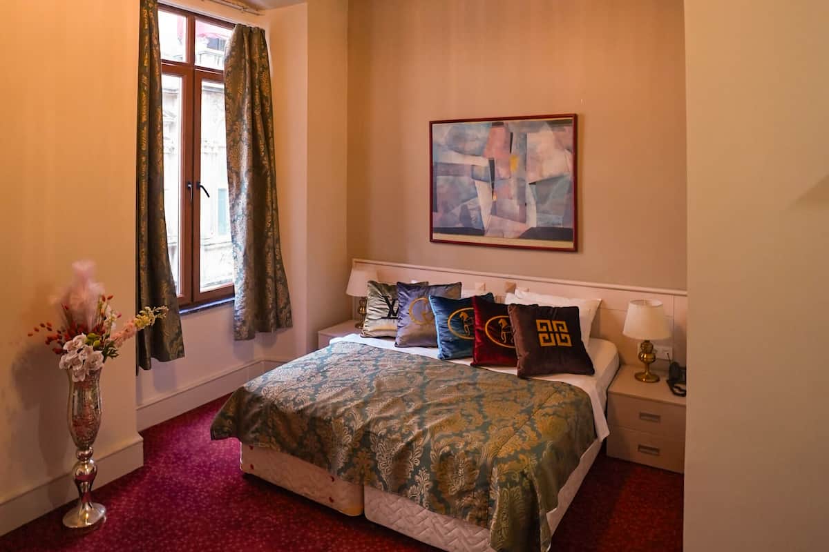 A bedroom with a bed, a nightstand, a lamp, a window with curtains, a painting on the wall, and a ceiling light fixture.
