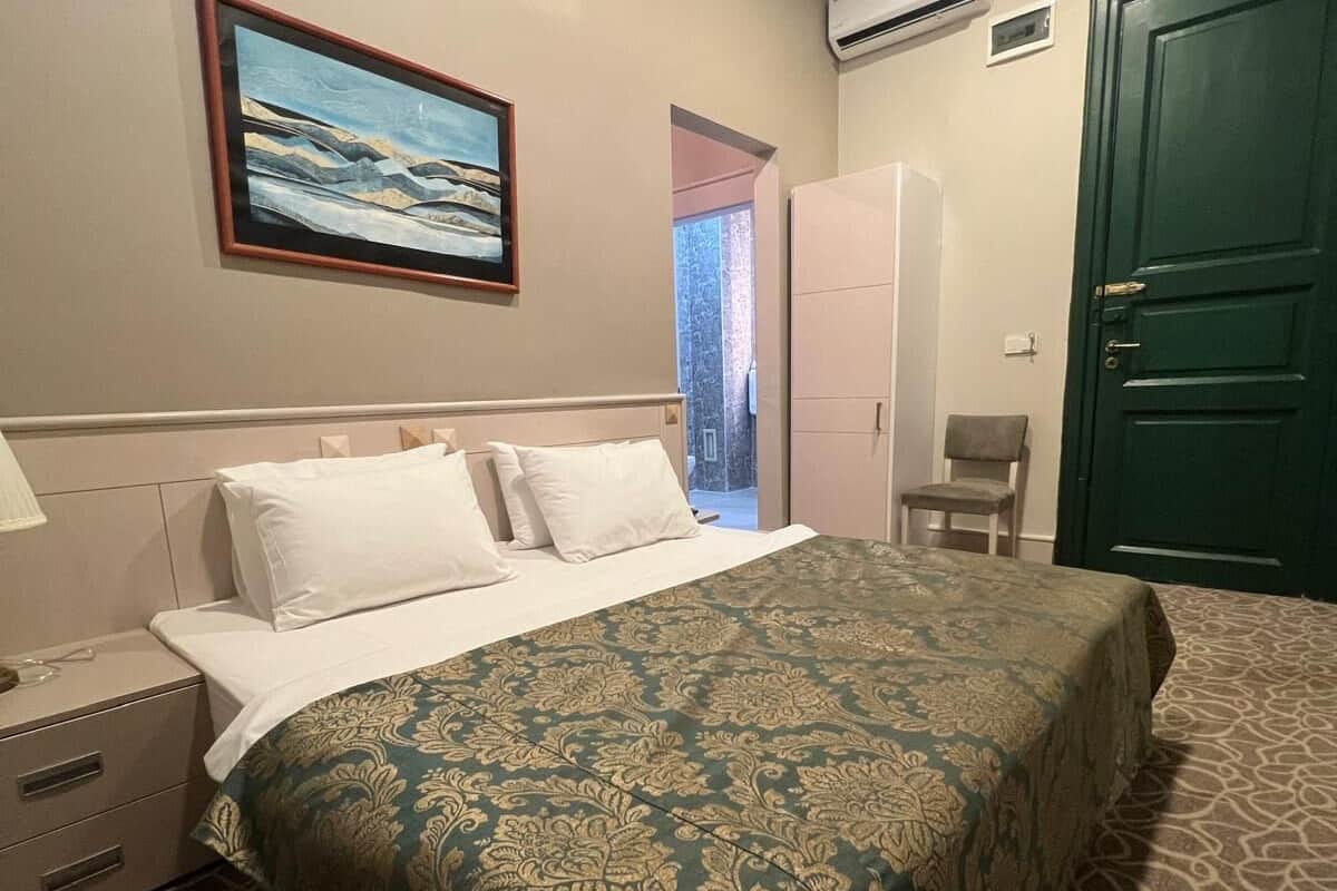 A hotel room with a bed, a chair, a wardrobe, and an air conditioning unit.