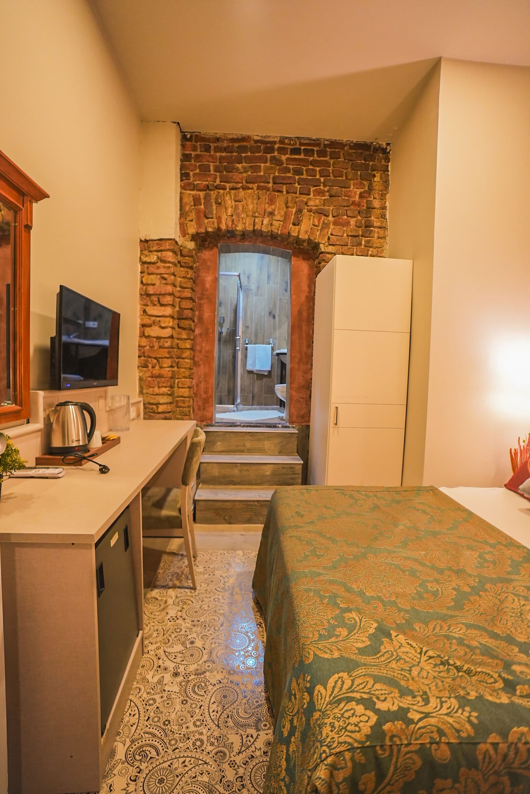Habitación de hotel con escritorio, cama con colcha estampada, televisor, tetera y pared de ladrillo.