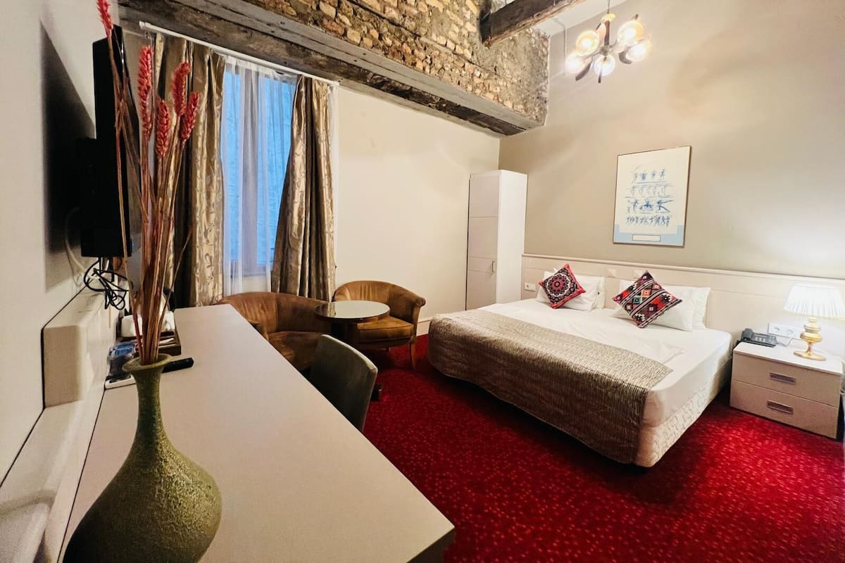 A hotel room with a bed, a desk, a chair, a TV, a vase, and a red carpet.