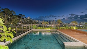 Una piscina rectangular con baldosas azules brillantes, rodeada por una terraza de madera y barandilla de vidrio, con vistas a un paisaje urbano que incluye edificios y montañas al fondo.