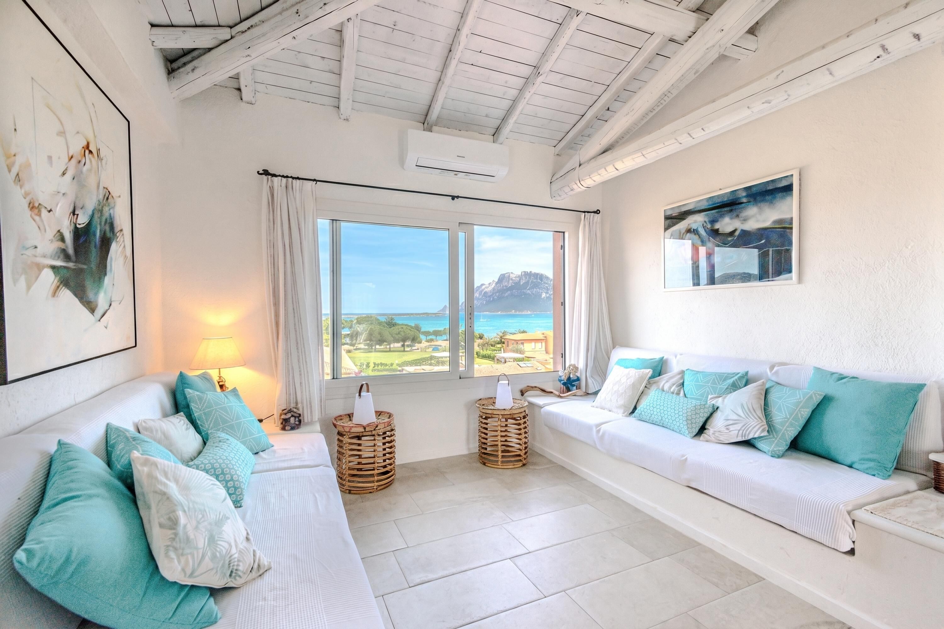 Una habitación con un sofá blanco, cojines color turquesa, cestas de mimbre y vistas al mar.