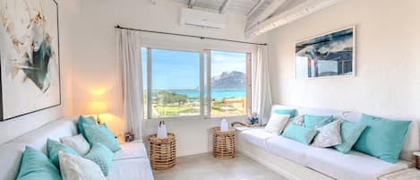 A room with a white sofa, teal cushions, wicker baskets, and a view of the sea.