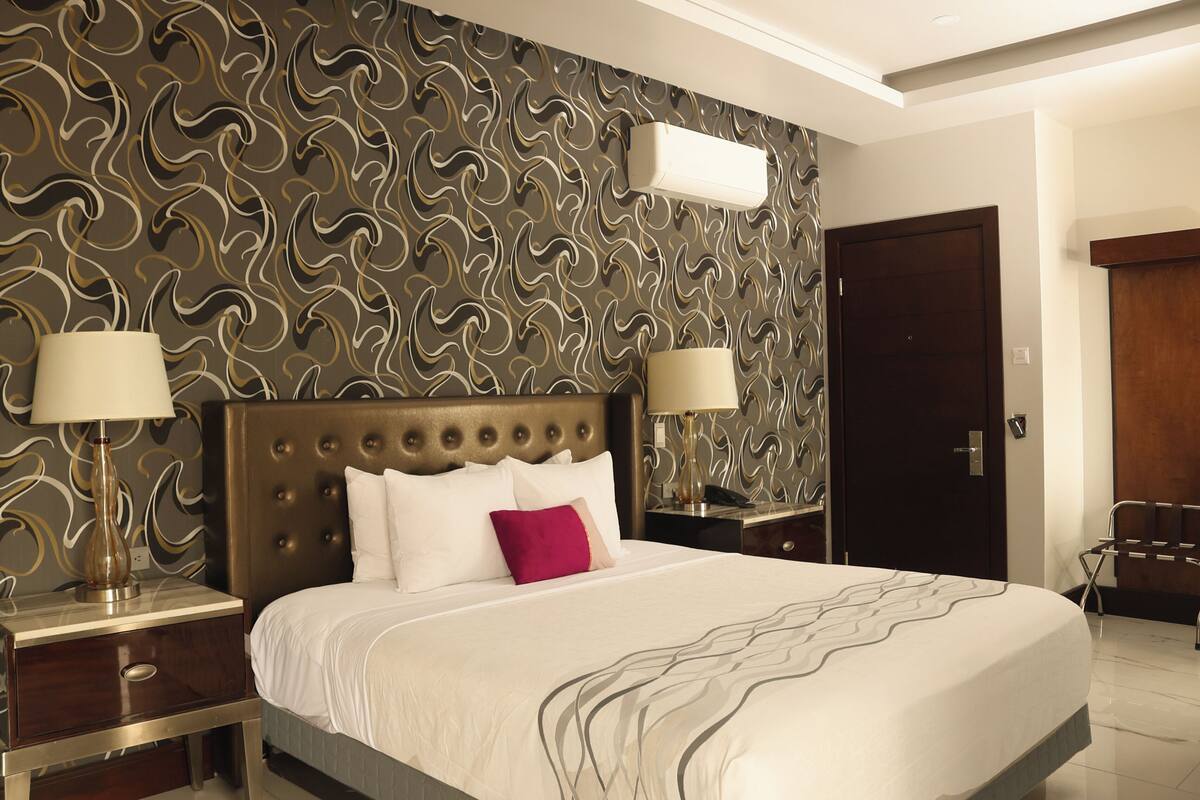 Ein Hotelzimmer mit einem großen Bett, zwei Nachttischen mit Lampen, einer dunklen Holztür und einem Wandteppich mit Muster.