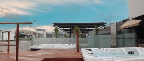 A rooftop terrace with hot tubs, wooden flooring, and a glass railing overlooking a cityscape.