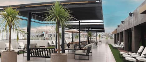 A rooftop lounge with outdoor seating, potted palm trees, and a view of the cityscape.
