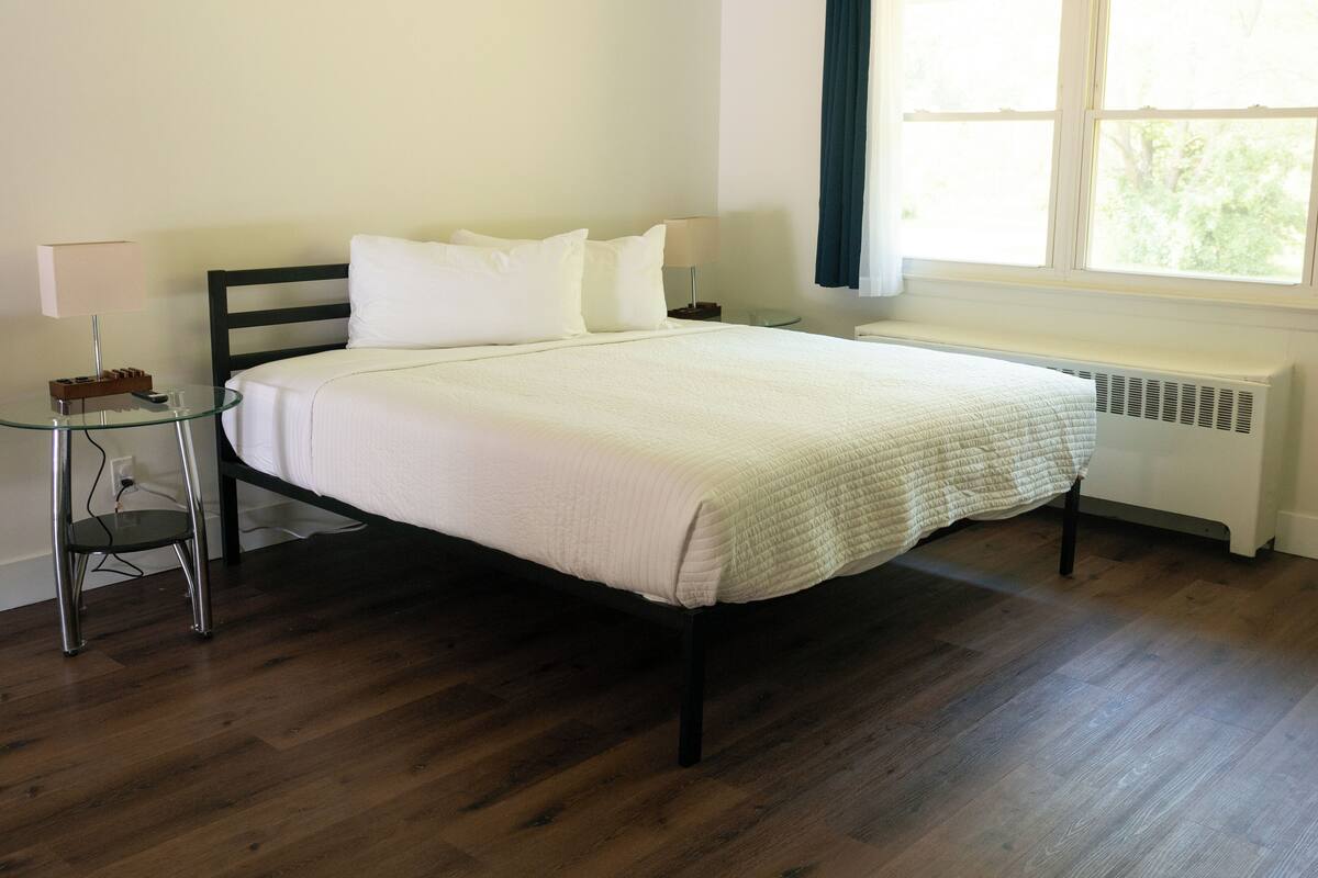 Una cama bien hecha con ropa blanca, una mesita de noche con una lámpara y una ventana con cortinas.