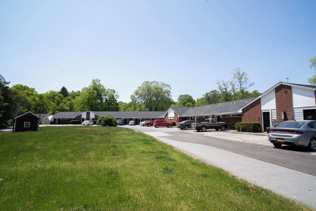 A parking lot with several cars, a small building, and a row of houses with a clear sky above.