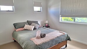 Un dormitorio con cama, almohadas, mesita de noche con lámpara y dos ventanas.