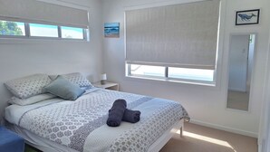Un dormitorio con una cama, almohadas, una mesita de noche con lámpara, una ventana con vistas y un cuadro en la pared.