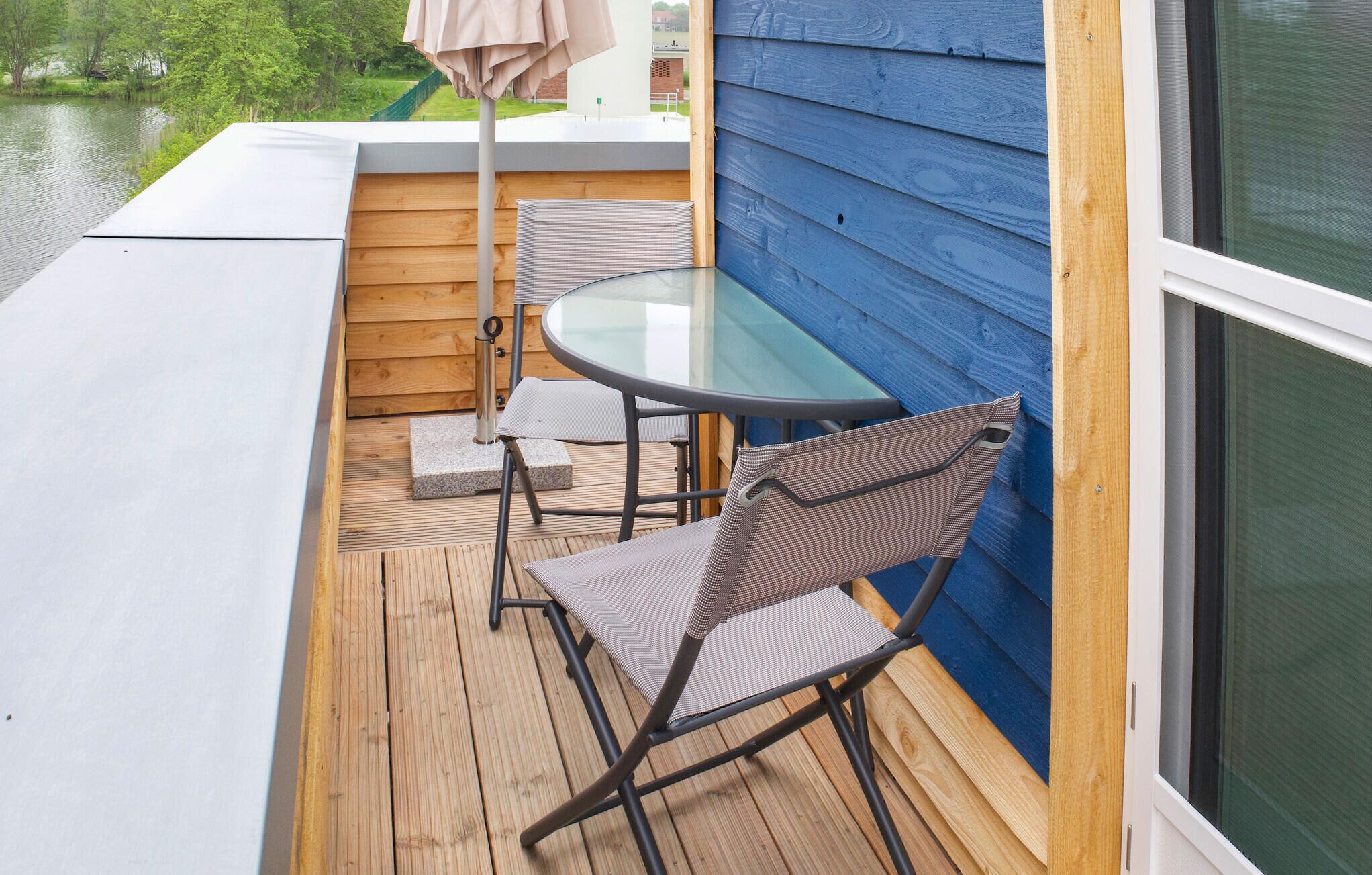 Une petite terrasse extérieure avec une table ronde en verre et deux chaises, un plancher en bois et un mur bleu.