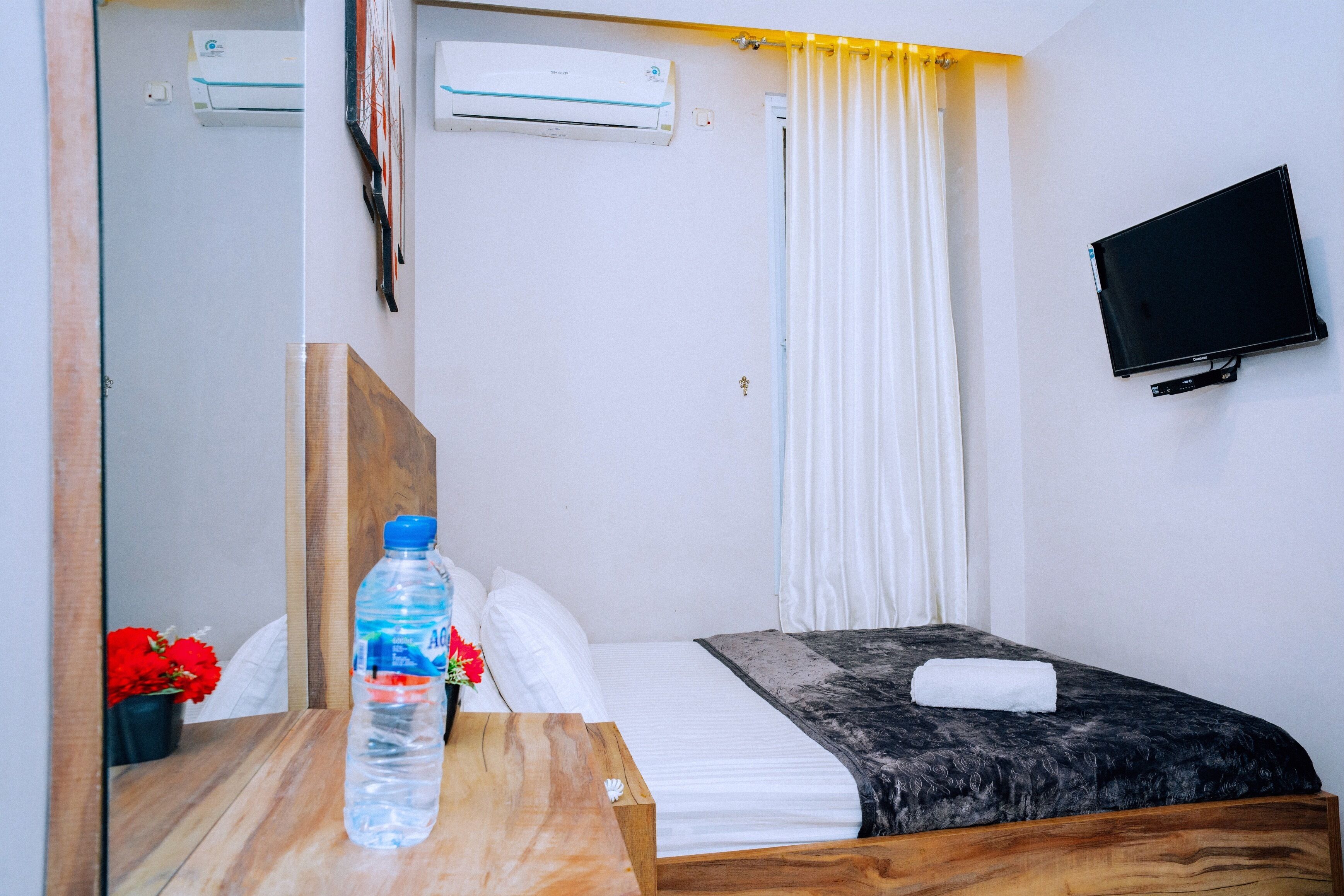 A hotel room with a bed, a wooden headboard, a water bottle, a TV mounted on the wall, and a window with curtains.