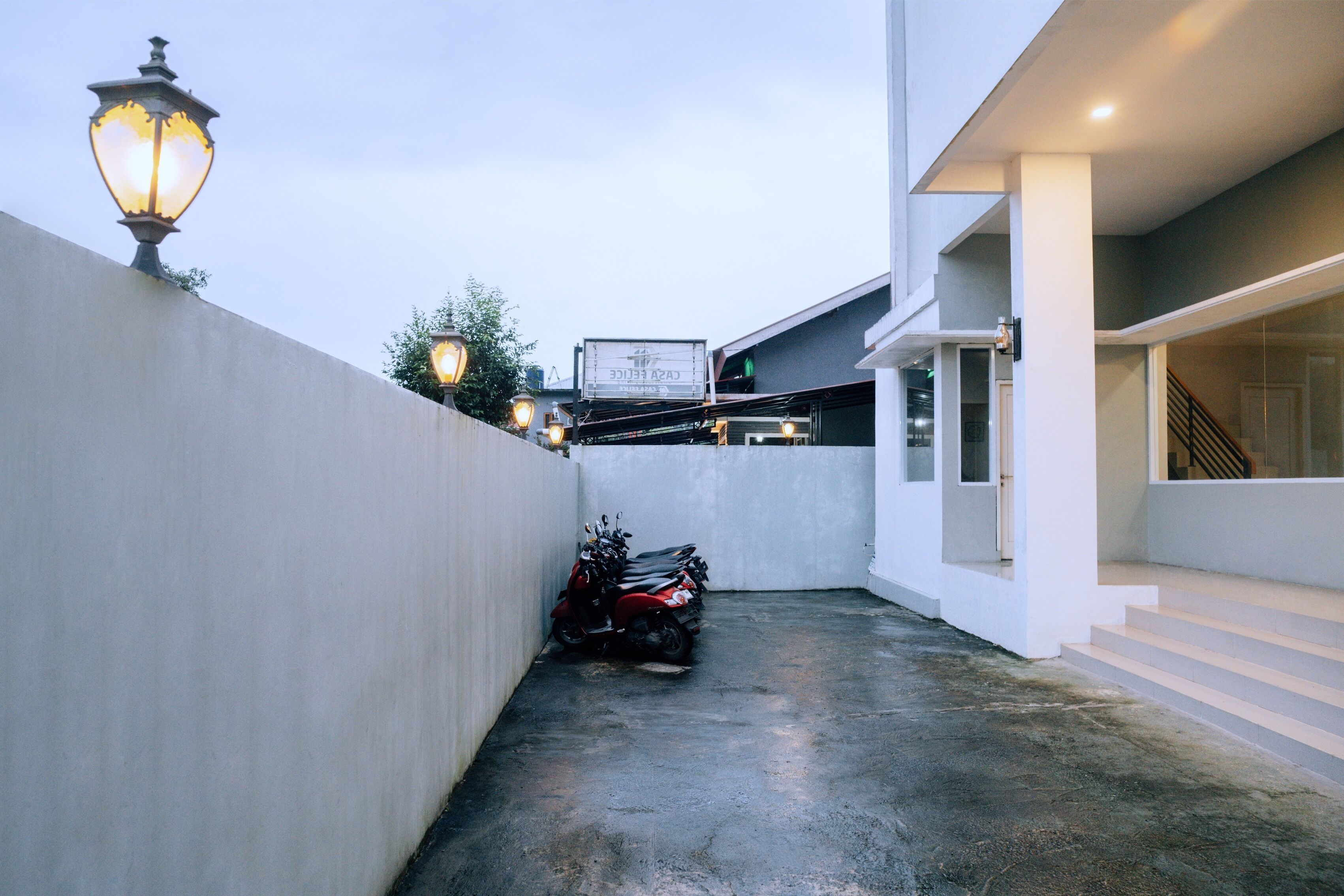 A narrow alleyway with a concrete floor, a white wall on one side, and a building entrance on the other. There are several motorcycles parked against the wall, and a street sign is visible in the background.