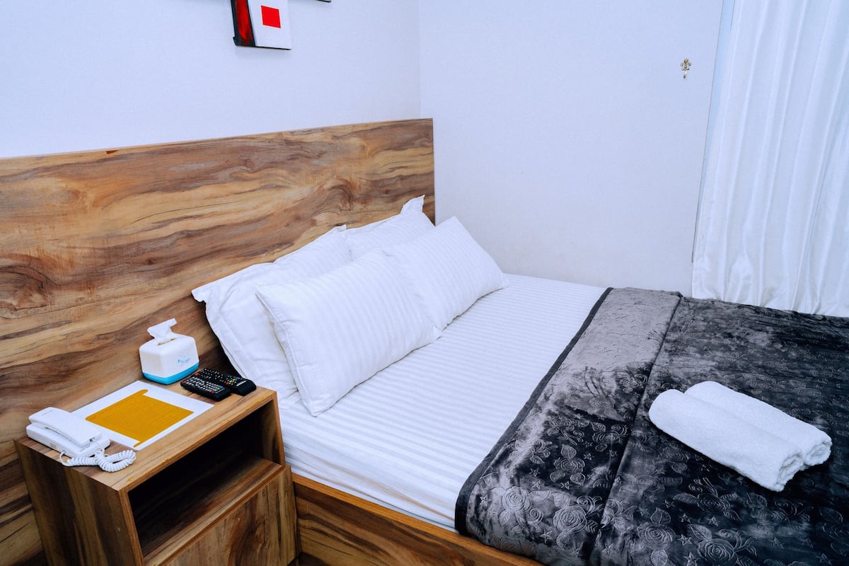 A neatly made bed with white linens, a wooden headboard, a bedside table with a tissue box, a remote control, and a folded towel.