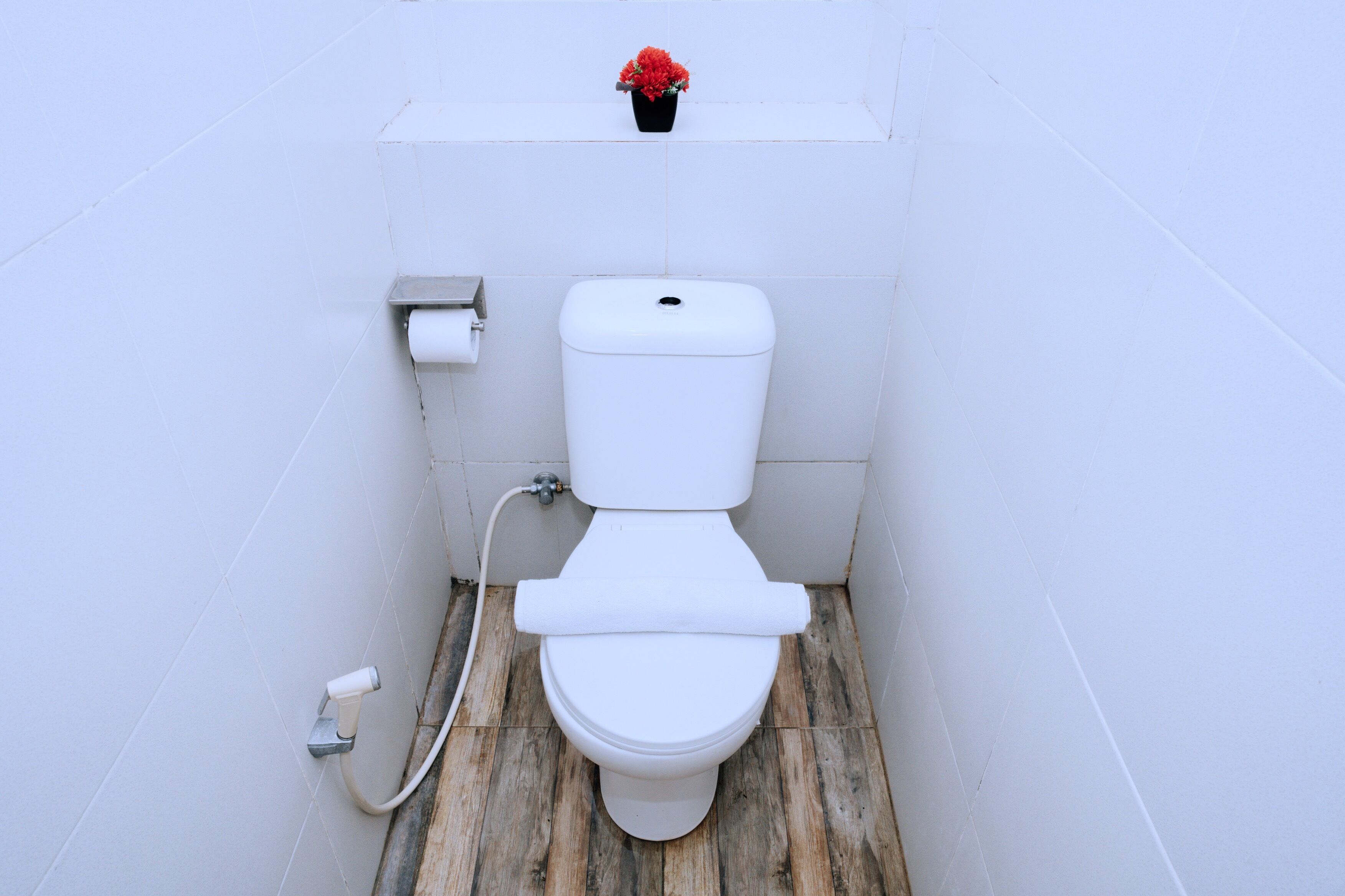 A toilet with a wooden floor, a toilet paper holder, and a small red flower on top of the tank.
