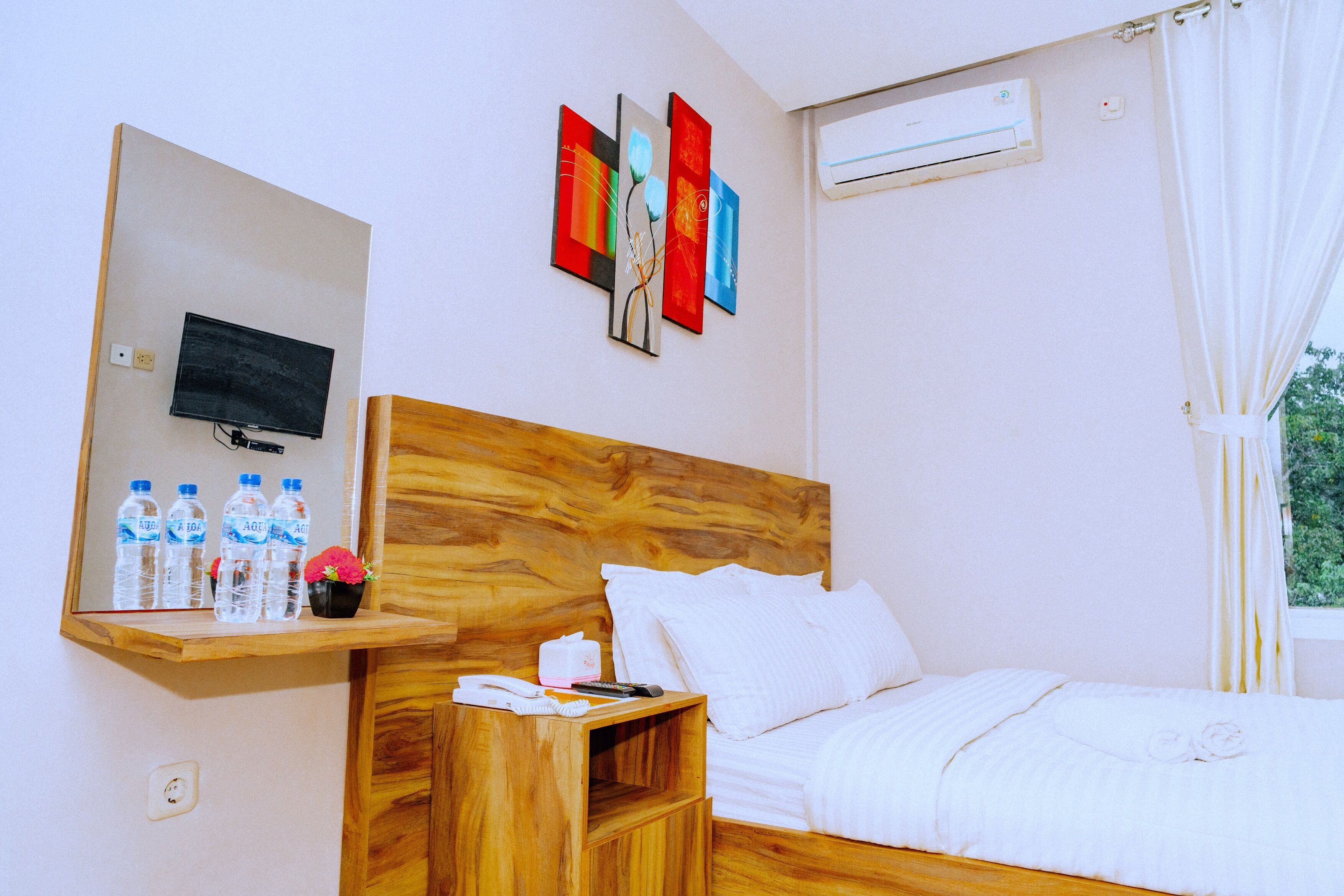 Habitación de hotel con cabecera de madera, cama con ropa de cama blanca, mesita de noche con un teléfono y botellas de agua, televisor de pantalla plana montado en la pared y ventana con cortinas.