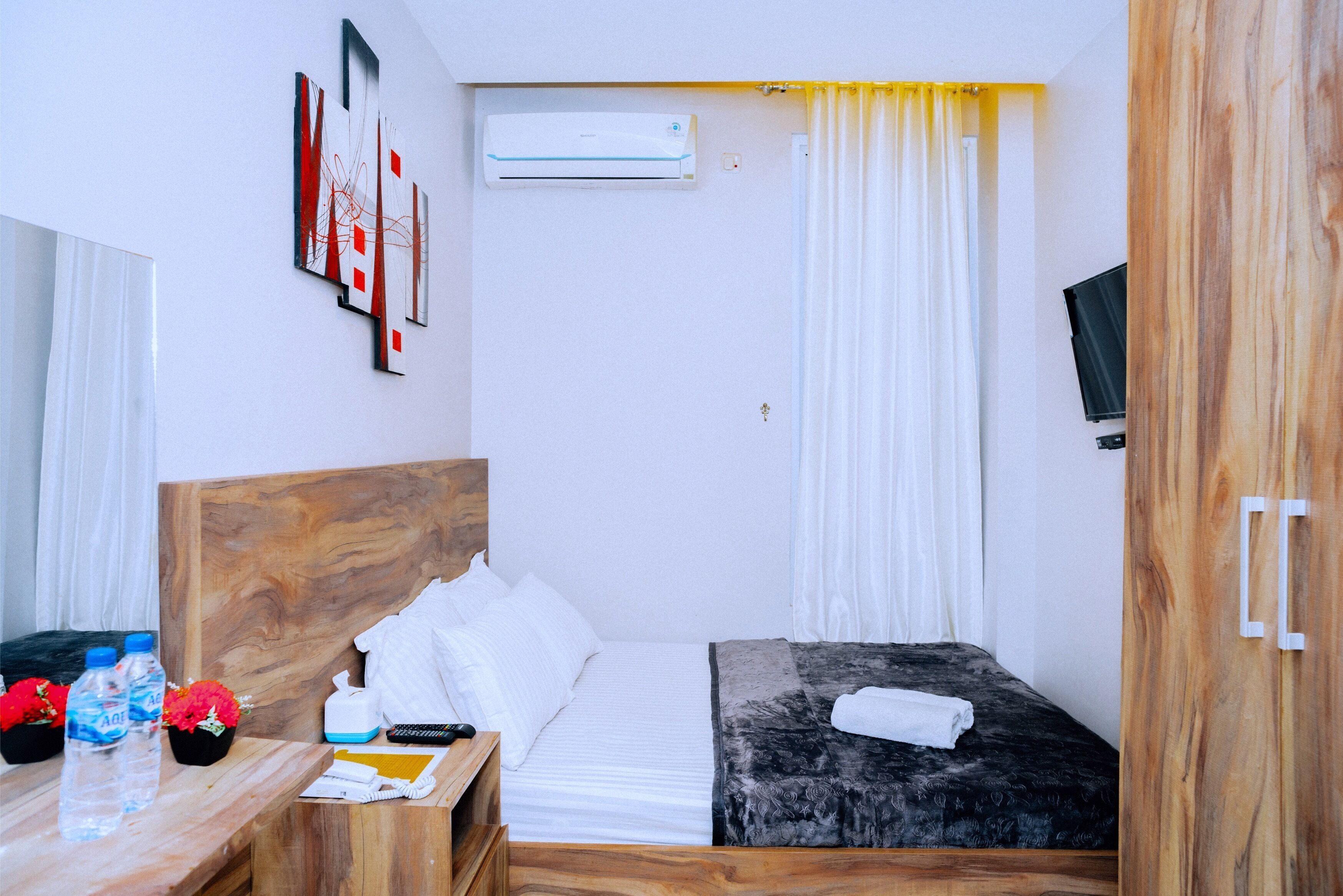 A hotel room with a wooden headboard, a bed with white and black bedding, a bedside table with a bottle and flowers, an air conditioning unit, and a TV on the wall.
