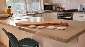 Cocina con armarios blancos, microondas, estufa y una isla grande con un rodillo. Hay dos sillas color turquesa y una ventana con persianas.
