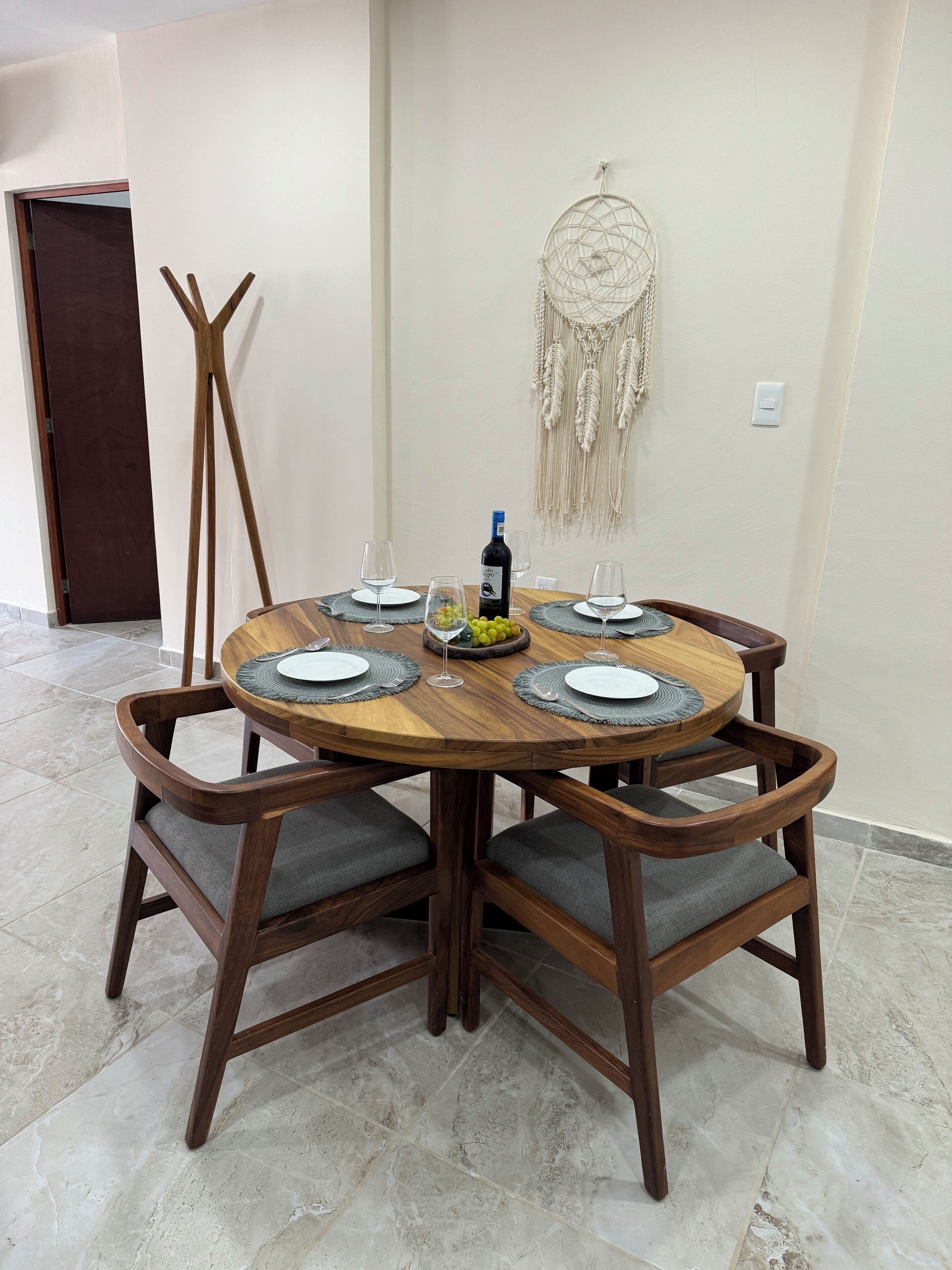 Una mesa de madera redonda con cuatro sillas, una botella de vino y un colgador decorativo de macramé.