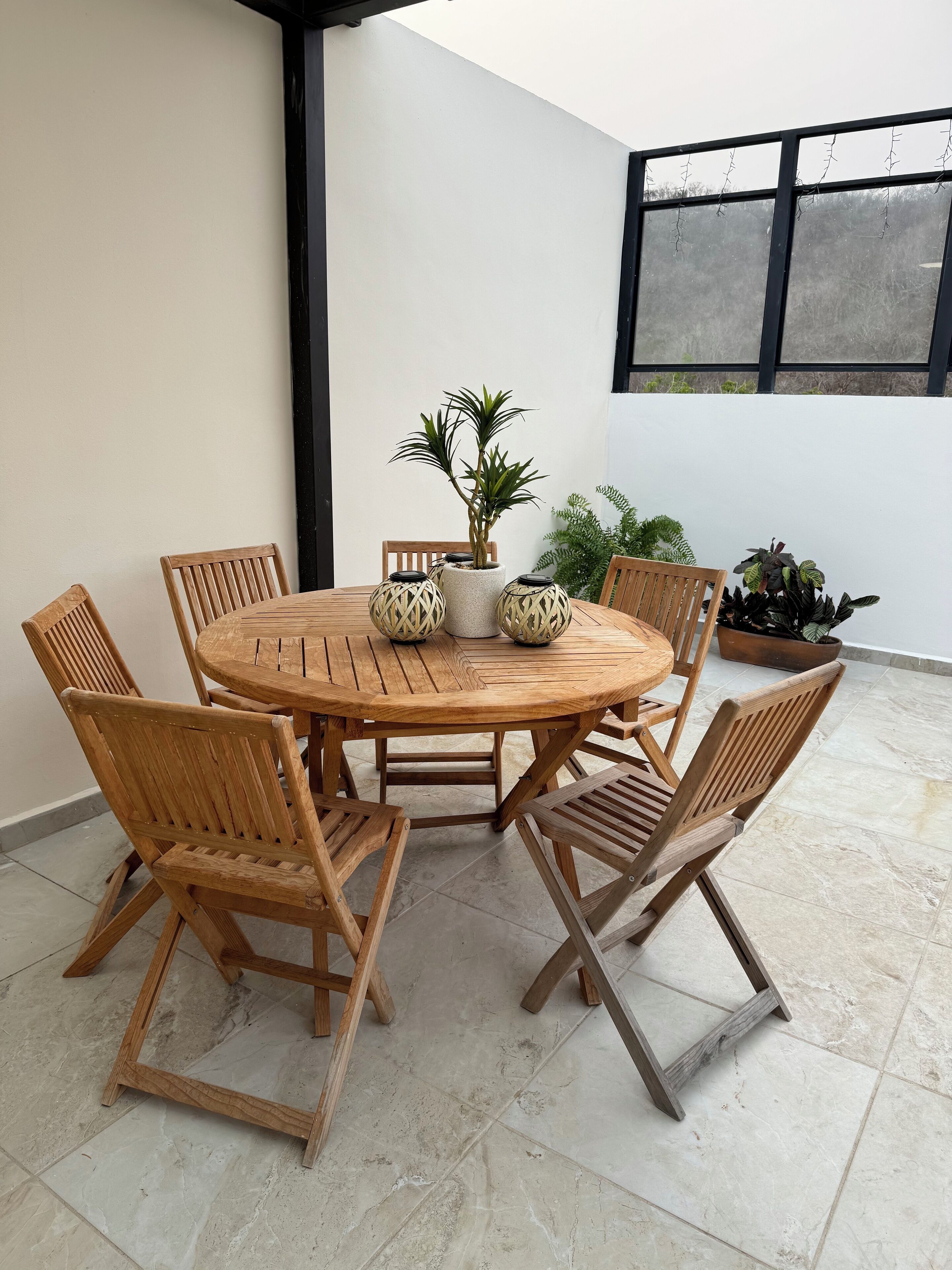 A round wooden table with six chairs, surrounded by potted plants, in a covered outdoor area with tiled flooring.