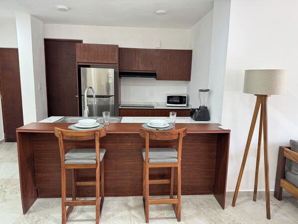 A modern kitchen with wooden cabinets, a stainless steel refrigerator, a microwave, and a blender on the counter. There are two wooden bar stools with cushioned seats and a small island with a sink and two glasses. - Nabani condominios T2 (Santa María Huatulco)