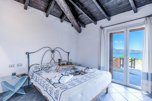 Ein Schlafzimmer mit Bett, Nachttisch, Tablett mit Gegenständen und Blick auf das Meer durch eine Schiebetür.