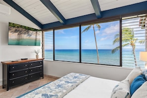 A bedroom with a large window, a bed, a dresser, and a painting of a beach scene.