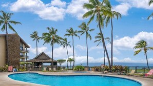 A swimming pool surrounded by palm trees and a building with a stone facade. - Rare LUX Oceanfront Views, Pool, Paradise - Papakea L402 (Lahaina)
