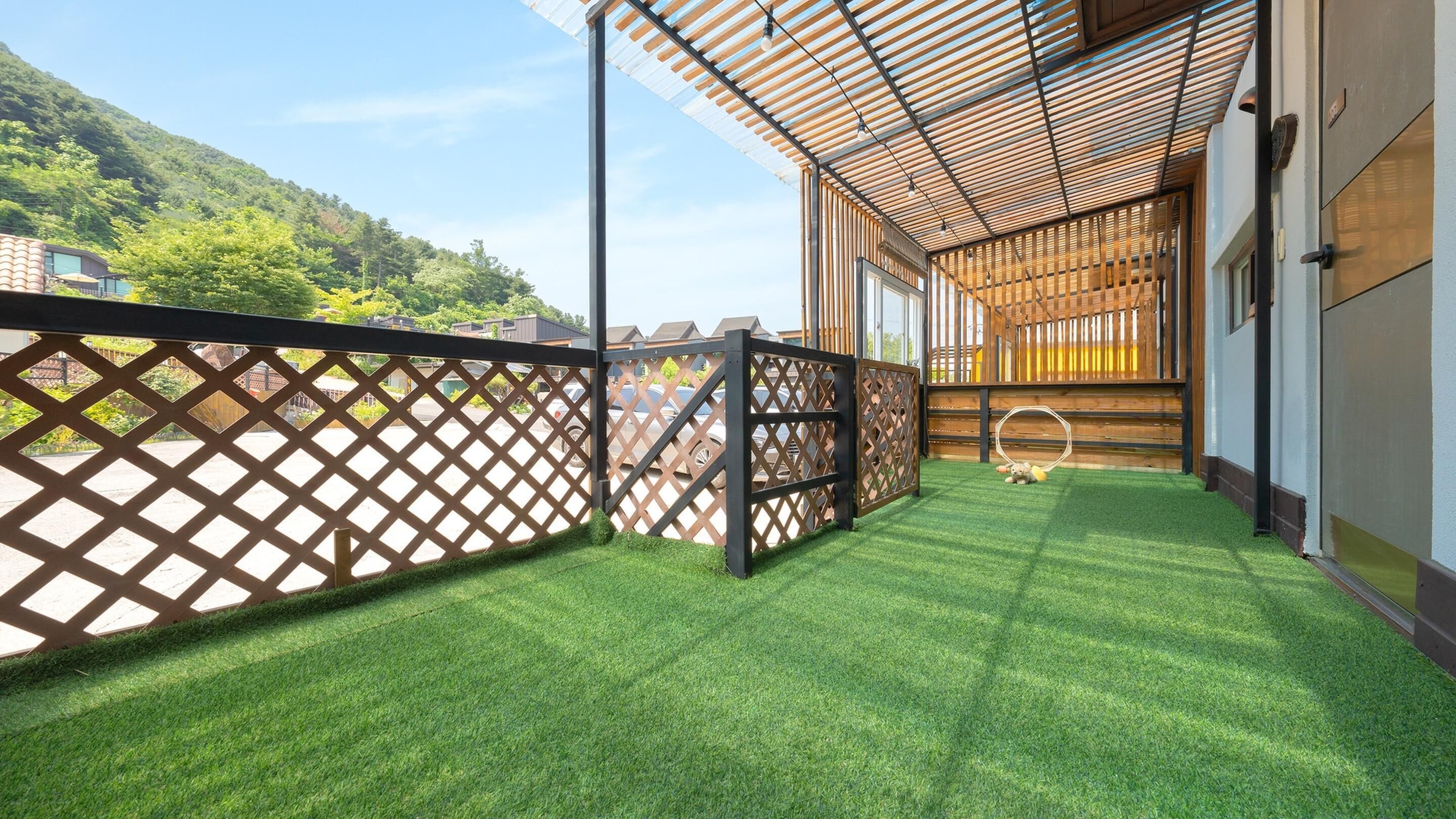 A covered outdoor area with artificial grass, a wooden fence, and a view of a hilly landscape.