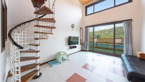 Un'ampia zona giorno con una scala a chiocciola in legno, una TV a schermo piatto, un divano e un'ampia porta scorrevole in vetro che dà su un balcone con vista sulle colline.