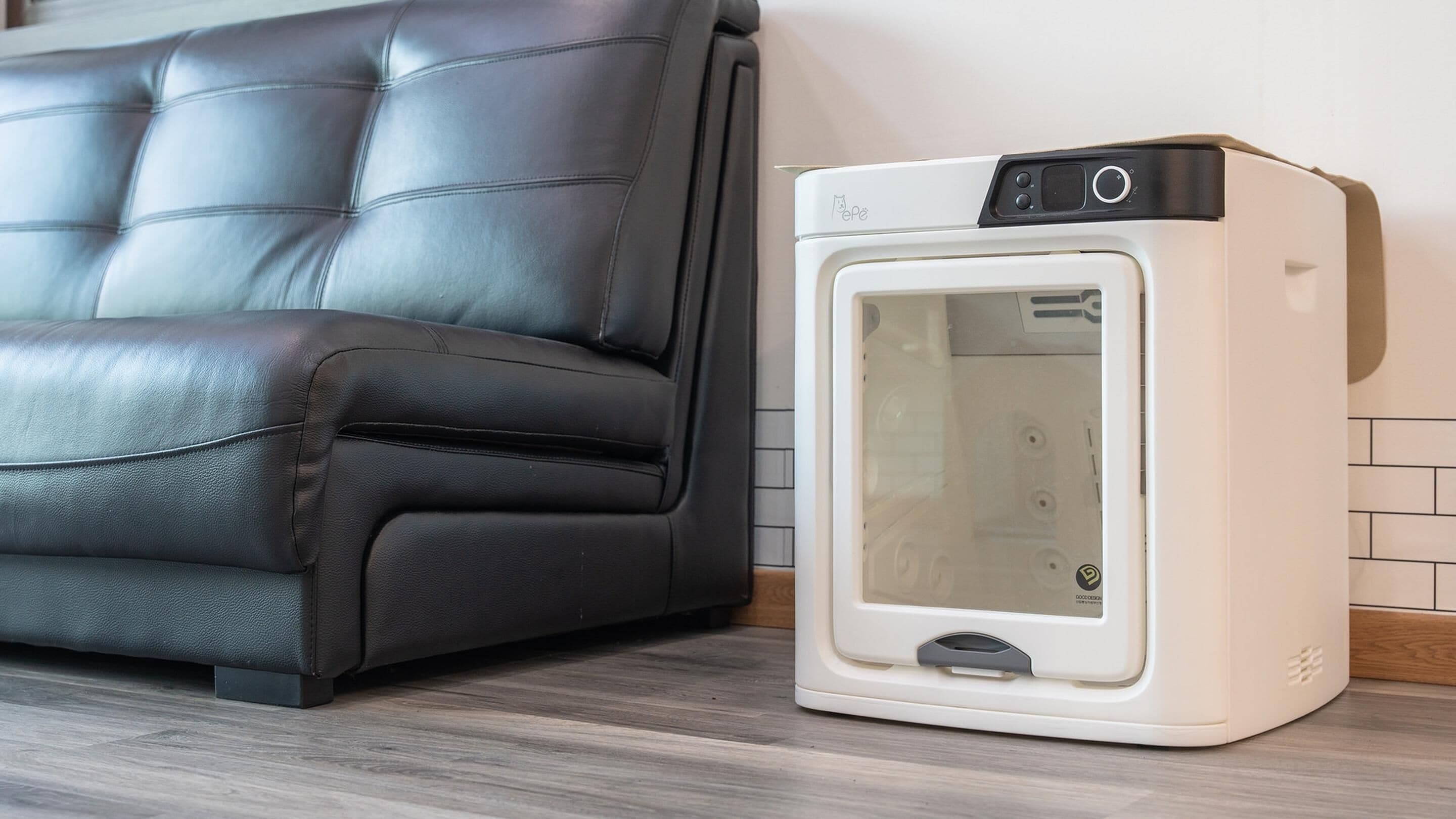 A white portable heater with a digital display and control buttons, placed on a wooden floor next to a dark brown leather sofa.