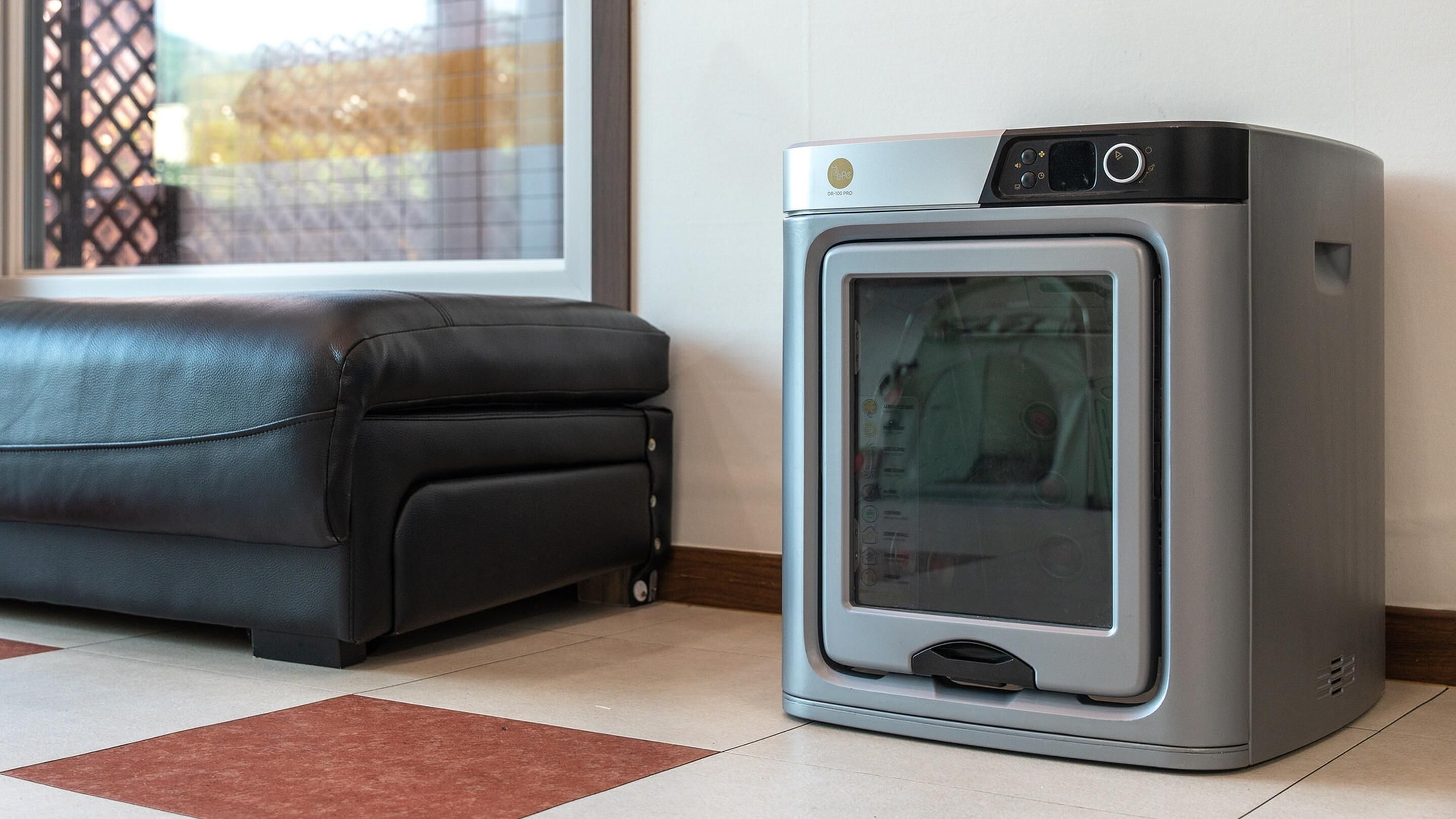 A compact, modern heater with a control panel, placed on a tiled floor next to a black leather sofa.