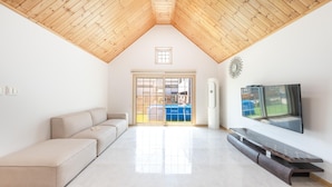 A spacious living room with a high wooden ceiling, a large window, a beige sofa, a flat-screen TV, and a built-in air conditioning unit. - Gapyeong Beilli pet Pool Villa Pension (Gapyeong)