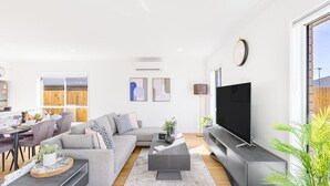 Een moderne woonkamer met een grijze bank, een salontafel, een flatscreen televisie en een eethoek met een houten tafel en stoelen.