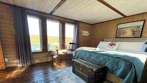 Un dormitorio de cabaña acogedor con una cama grande, piso de madera rústica, una mesita con una silla y un ventanal con vistas a un paisaje pintoresco.