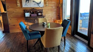 Interior de madera con una mesa redonda, sillas azules y beige, un cuadro de ballenas y vistas a una parrilla exterior.