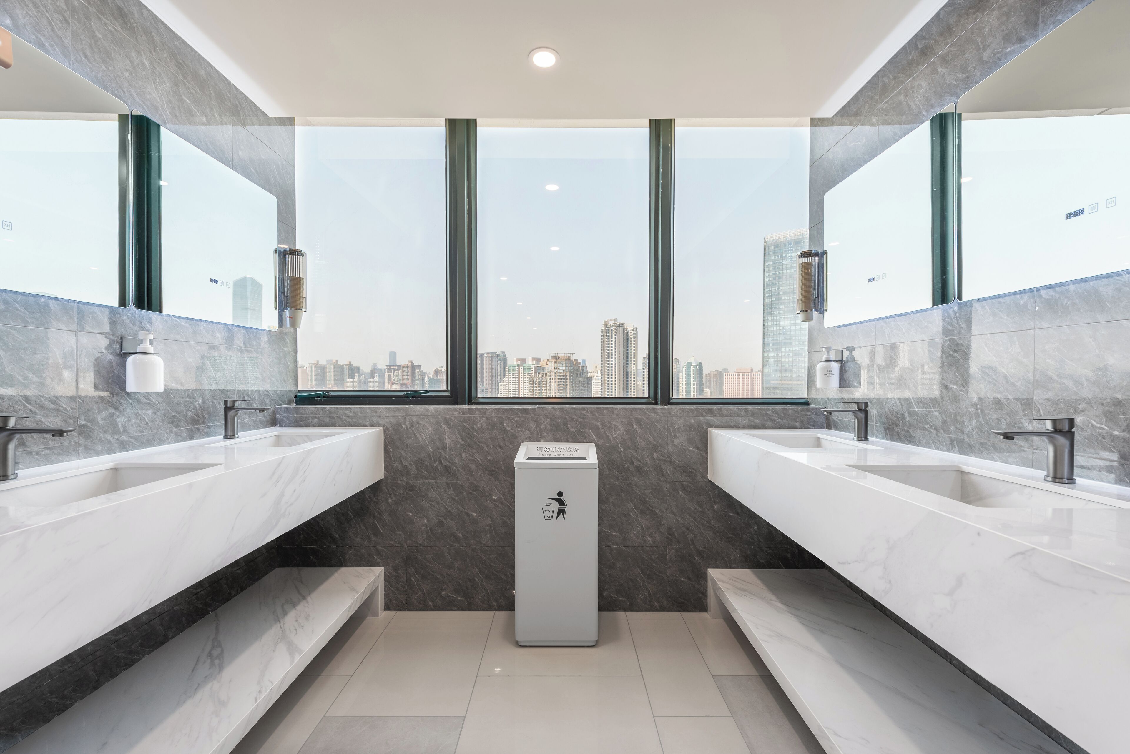 A modern bathroom with two sinks, a trash bin, and a city view through large windows.