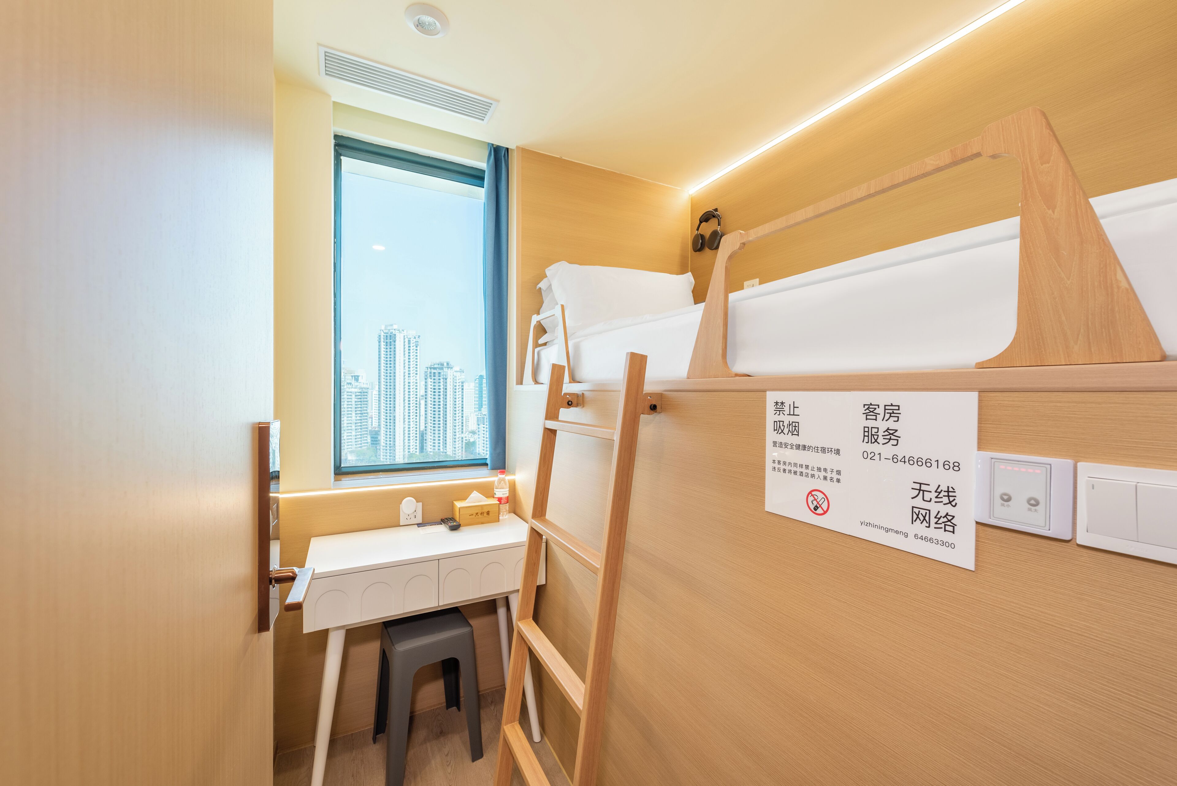 A hotel room with a desk, a window with a city view, a bed with a wooden headboard, and a sign with contact information.
