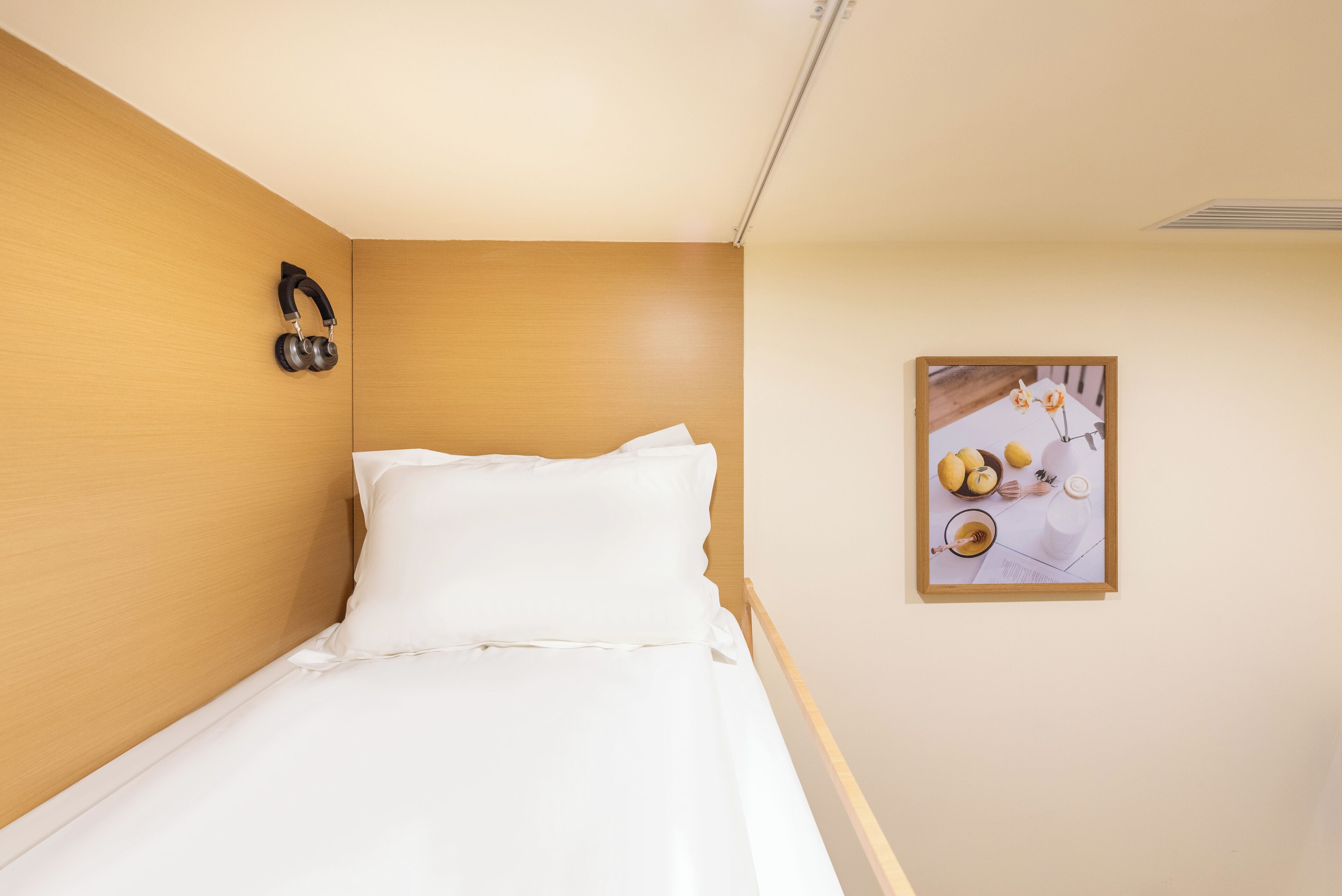 A neatly made bed with white linens, a pair of headphones on the wall, and a framed picture of lemons and honey on the wall.