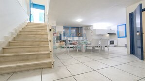 Una zona d'estar àmplia amb una escala, una zona de menjador amb mobles blancs i una cuina amb detalls blaus.
