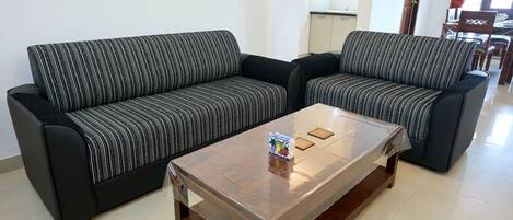 A living room with two black sofas with white stripes, a glass coffee table, and a kitchen area in the background.